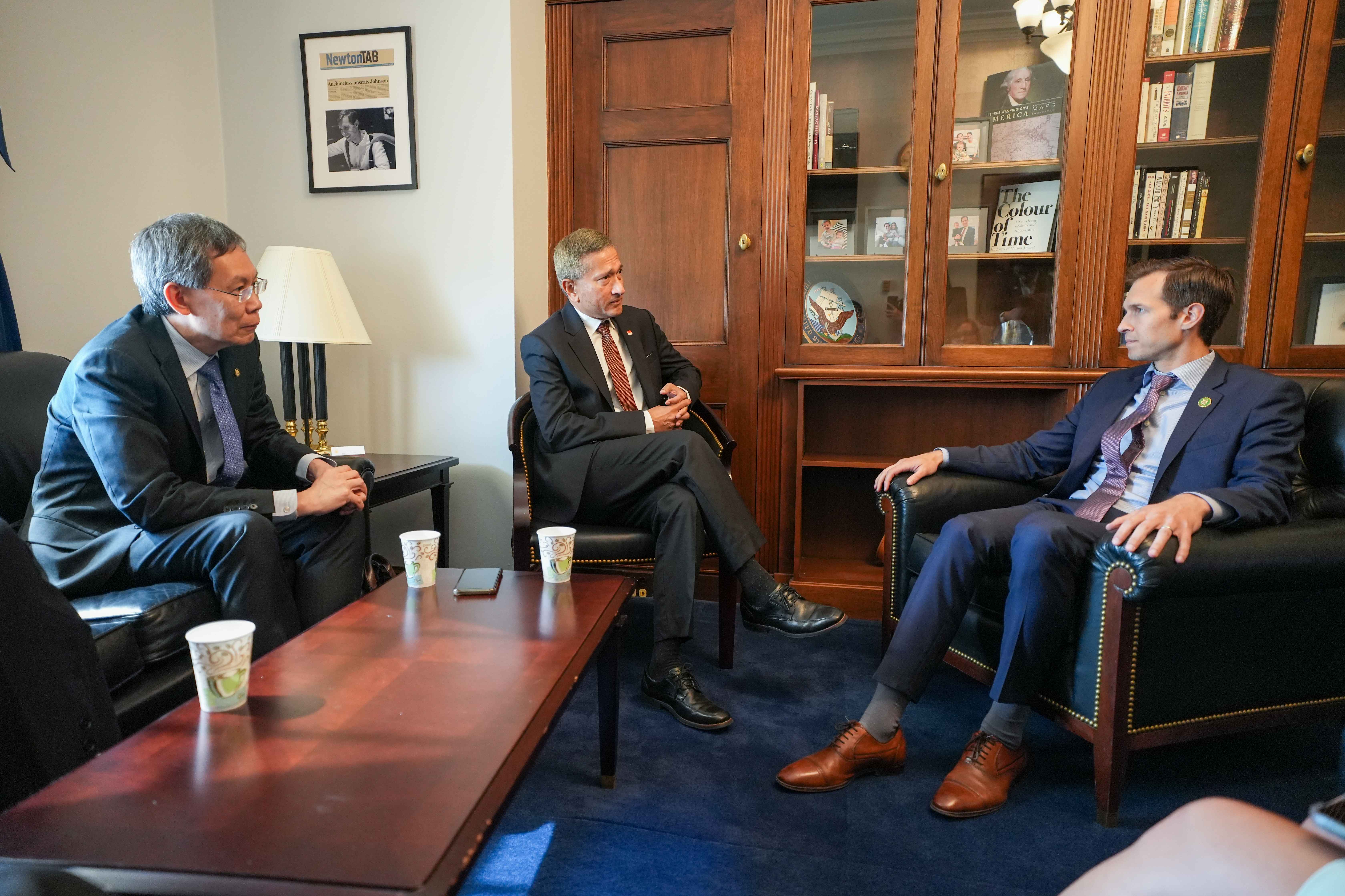 Three men in suits sit in armchairs in an office, talking near a wooden table with paper cups.